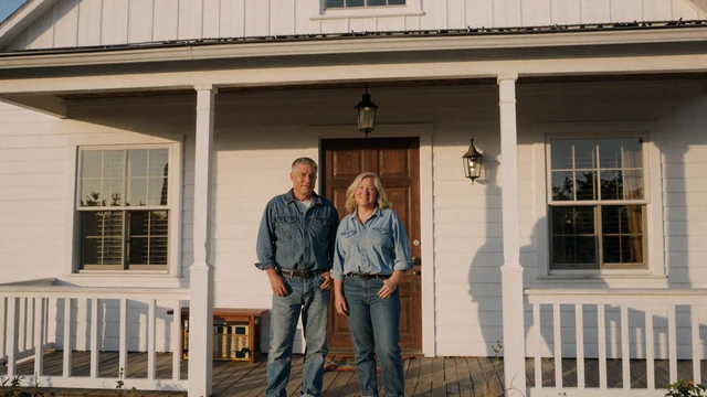 Mature farmer couple on the porch of a white farmhouse at golden hour, farmer dating site for rural singles 50+